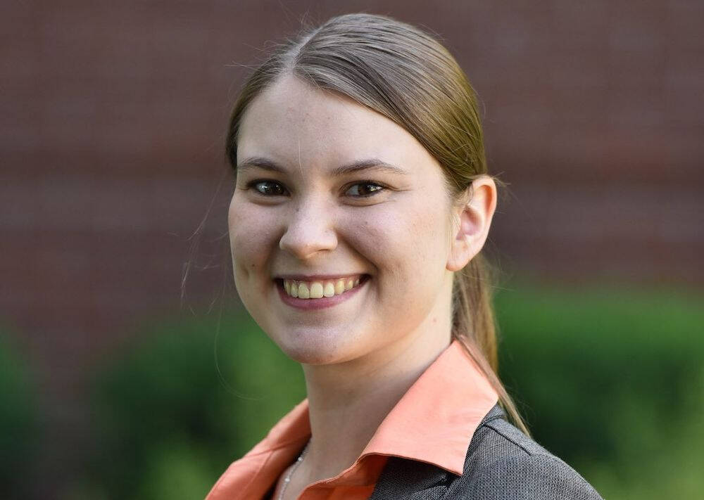 A smiling woman with straight brown hair tied back, wearing a gray blazer over an orange collared shirt, standing outdoors in front of a brick wall and greenery